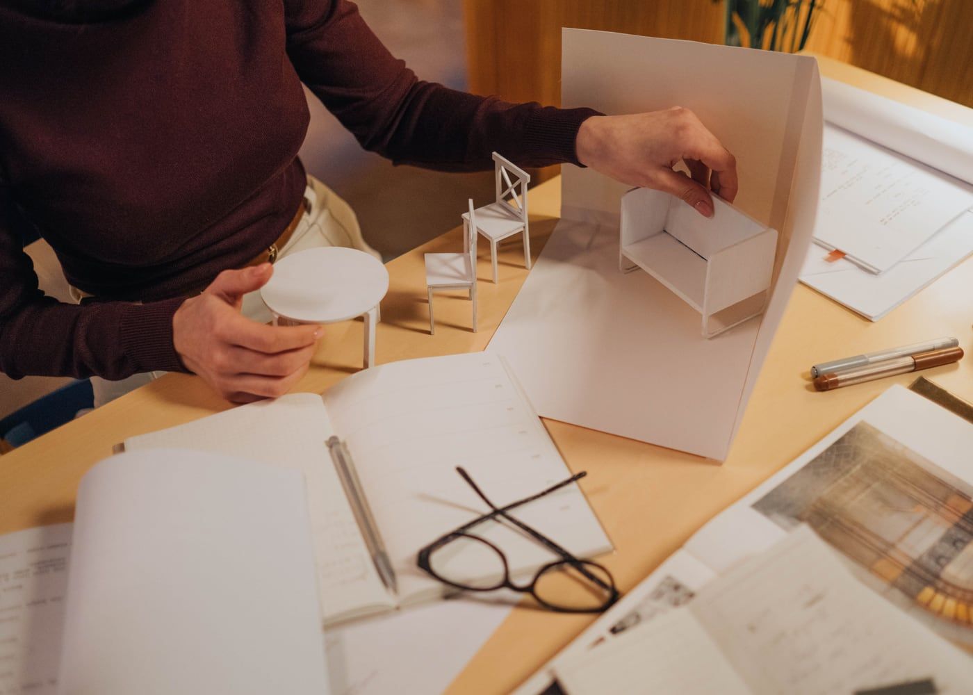 Hände arbeiten an einem weißen Architekturmodell, daneben Zeichnungen und Werkzeug auf dem Tisch